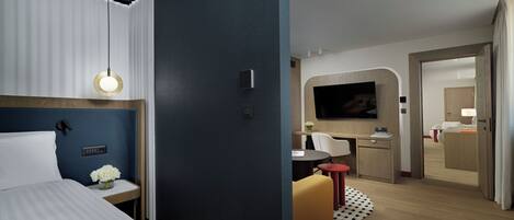 Habitación de hotel moderna con cama, mesita de noche y escritorio con silla. Hay un televisor montado en la pared y una puerta que da a otra habitación.