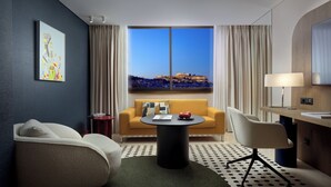 A hotel room with a sofa, a round table with a book and apples, a chair, a lamp, and a view of a historic building.