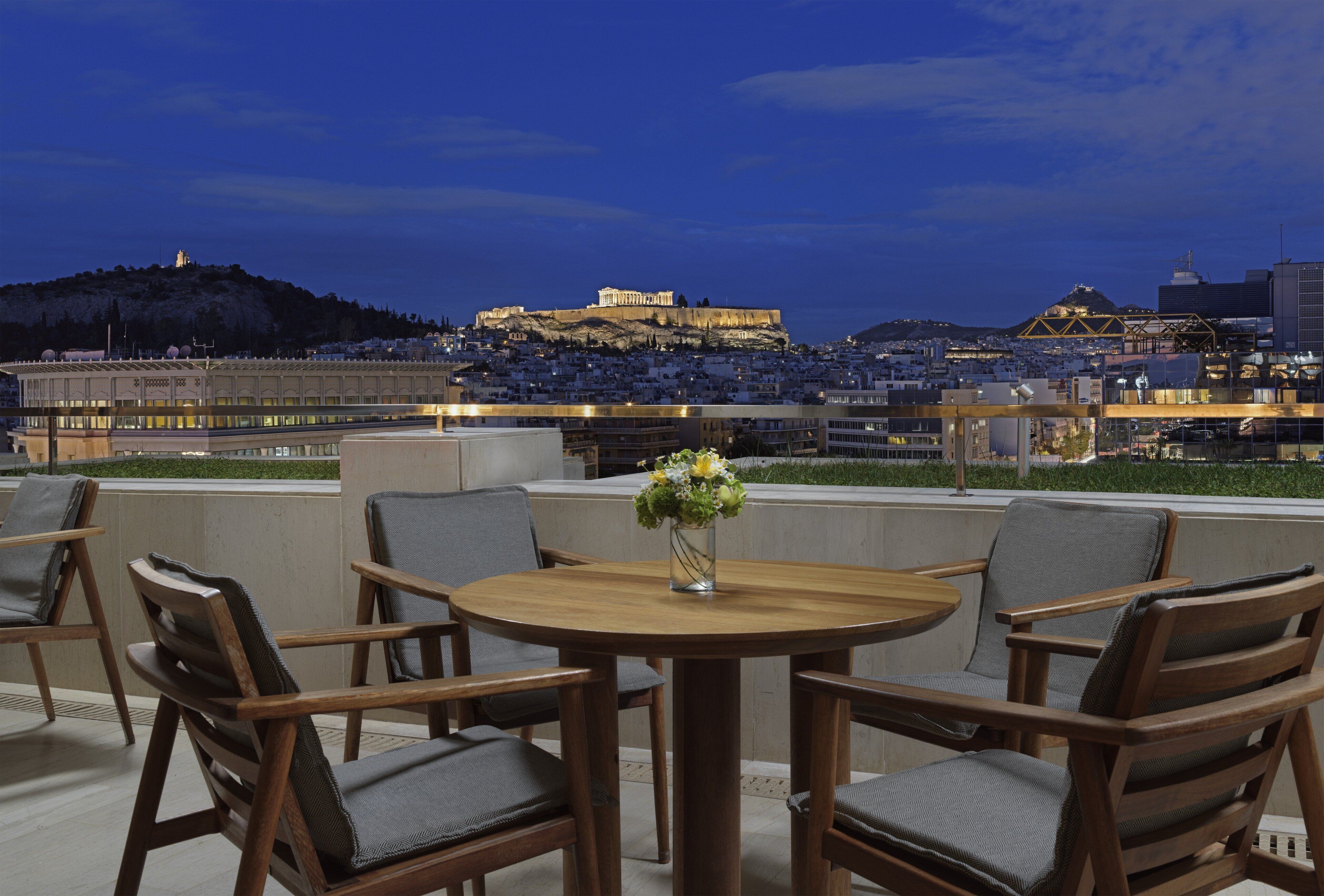 A rooftop terrace with a round wooden table and six chairs, overlooking a cityscape with a prominent hill and a large building.