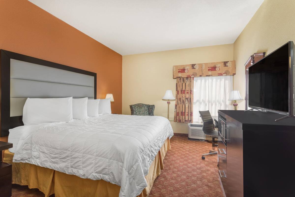 A hotel room with a large bed, a desk with a flat-screen TV, a chair, and a lamp.