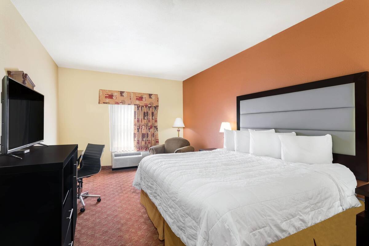 A hotel room with a bed, a desk with a computer, a chair, a lamp, and a window with curtains.
