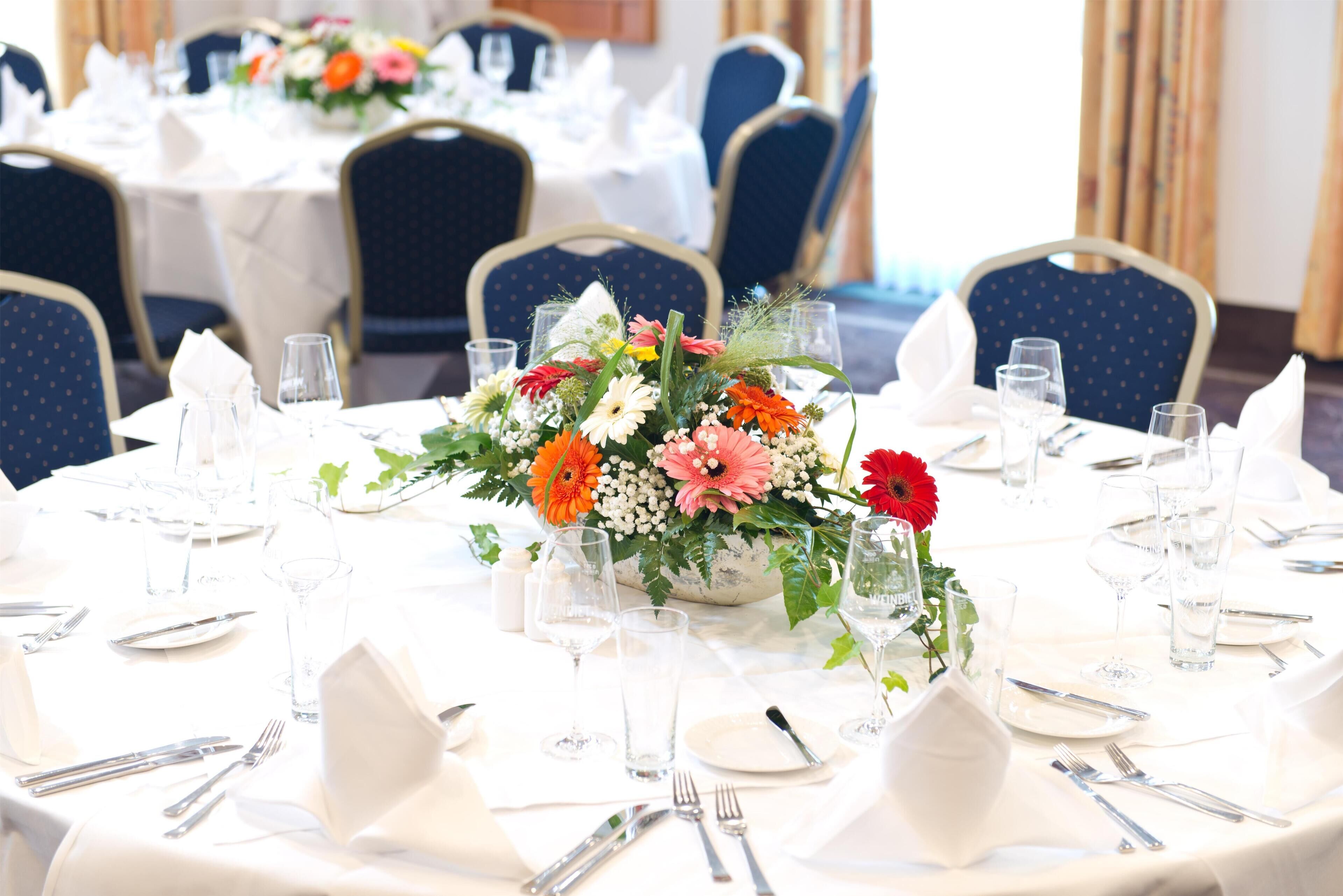 A round dining table set for a formal event with white tablecloths, neatly arranged silverware, and a centerpiece of assorted flowers.