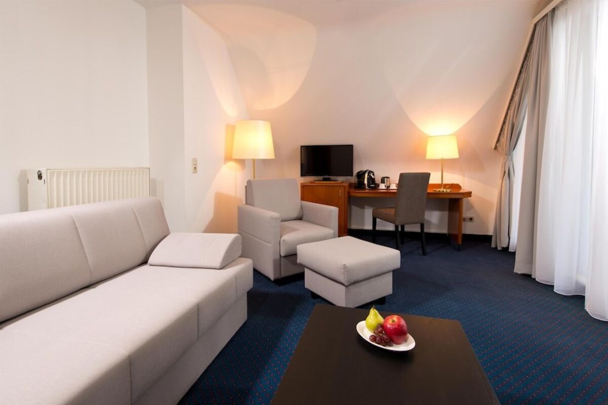 Une chambre d’hôtel avec un canapé, deux fauteuils, une table basse avec des fruits, un bureau avec une télévision, des lampes et un radiateur.