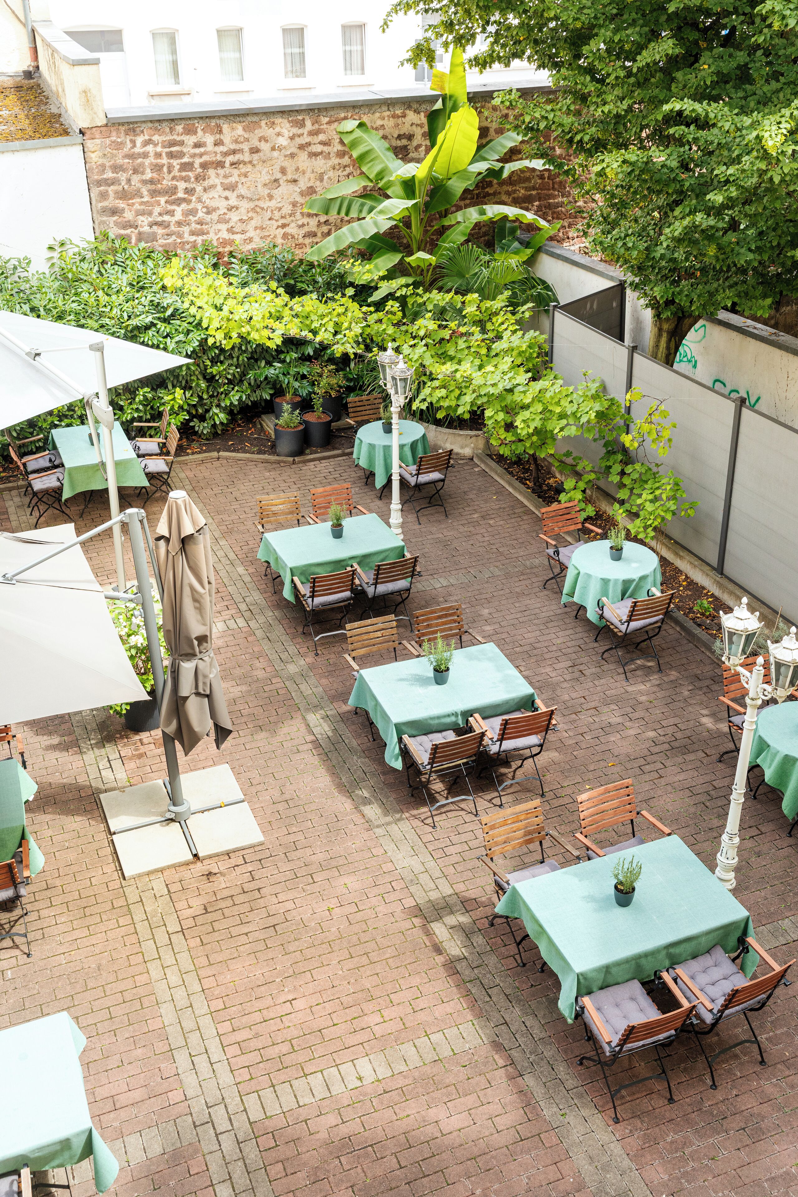 An outdoor patio area with multiple tables and chairs, surrounded by lush greenery and brick flooring.
