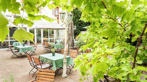 An outdoor patio area with tables and chairs, covered by umbrellas, and surrounded by greenery. - ACHAT Hotel Neustadt an der Weinstraße (Neustadt an der Weinstrasse)