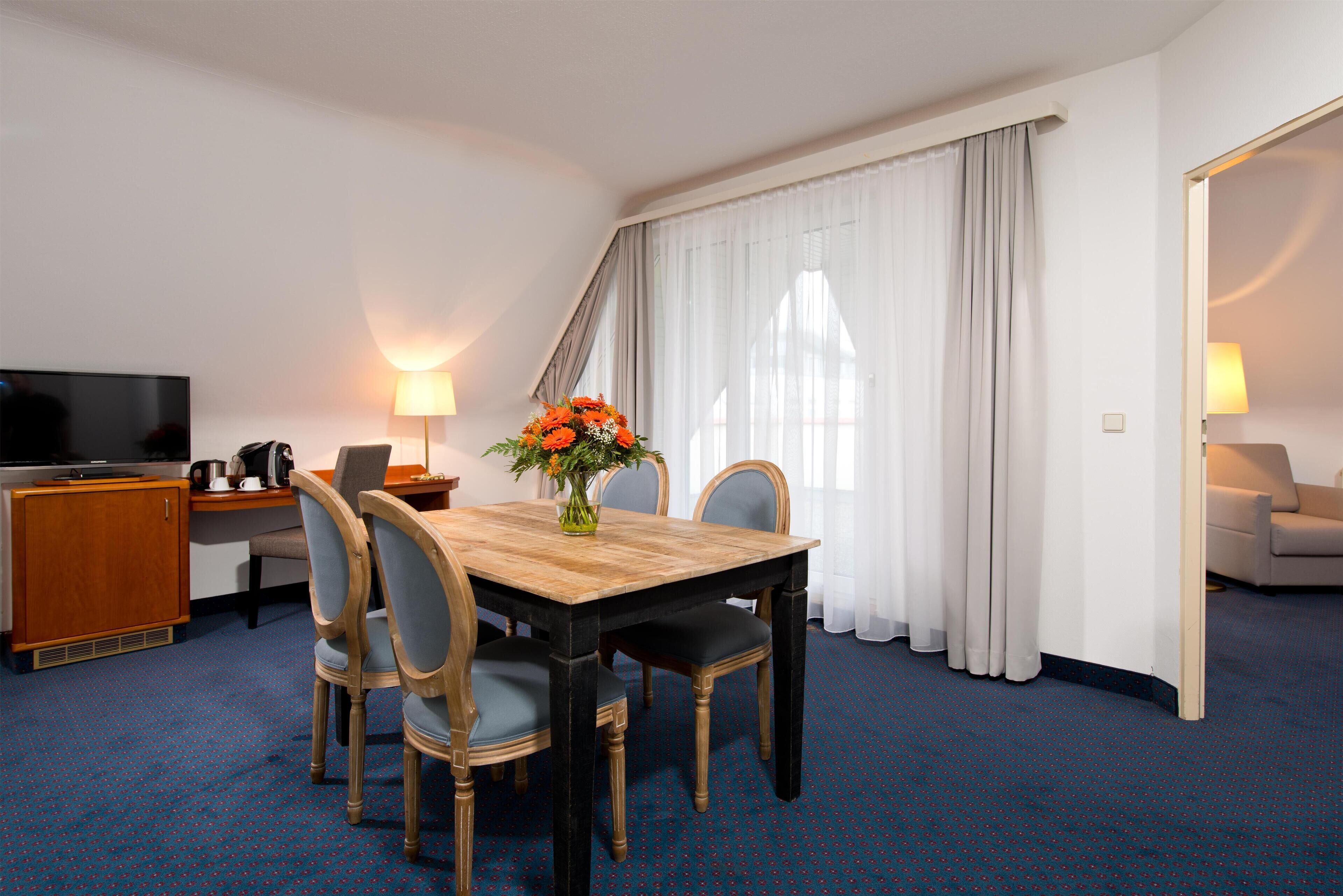 A hotel room with a wooden dining table, four chairs, a TV, a desk with a lamp, and a flower vase.