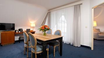 A hotel room with a wooden dining table, four chairs, a TV, a desk with a lamp, and a flower vase.