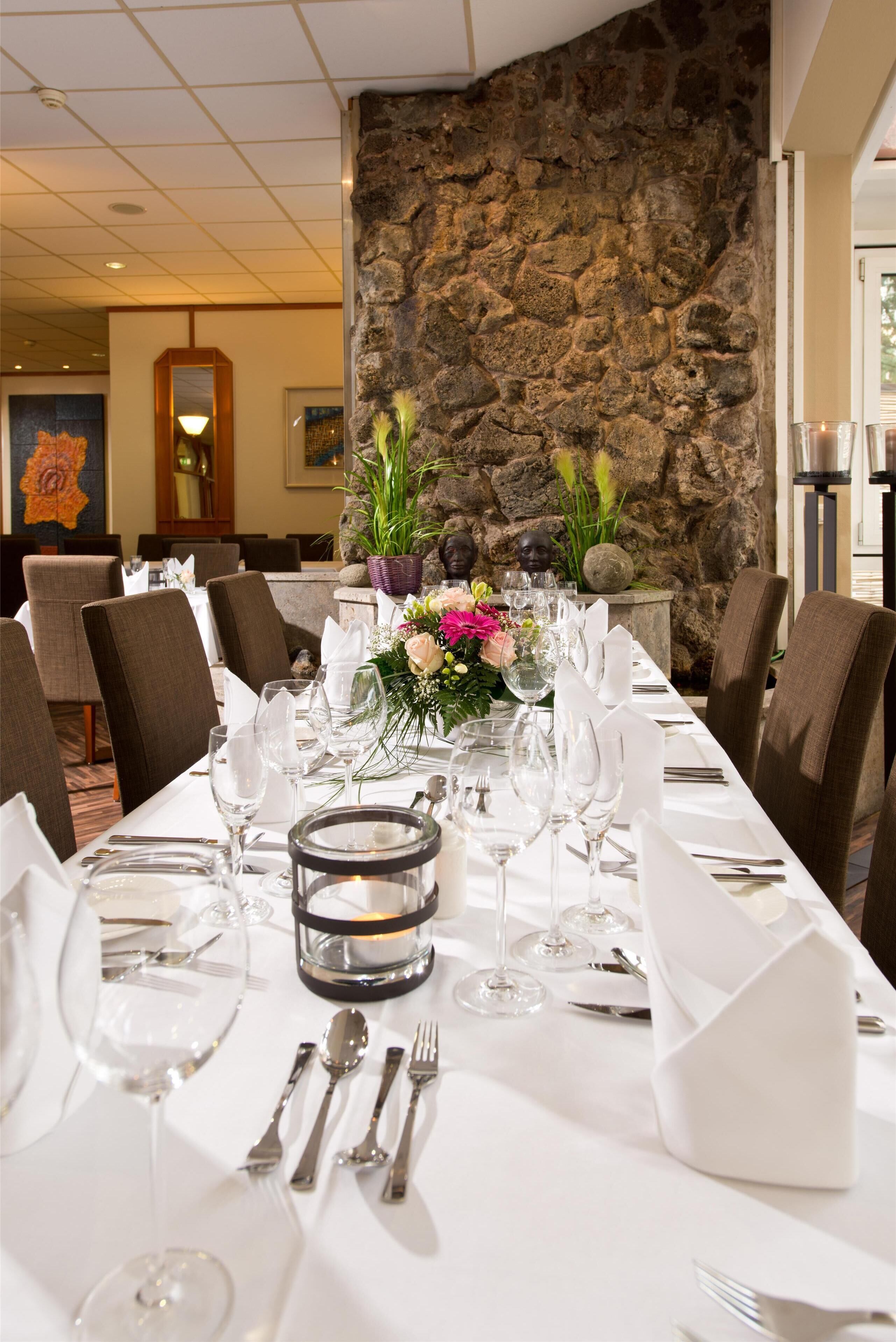 A long dining table set for a formal meal in a hotel room with a stone wall and a painting on the wall.