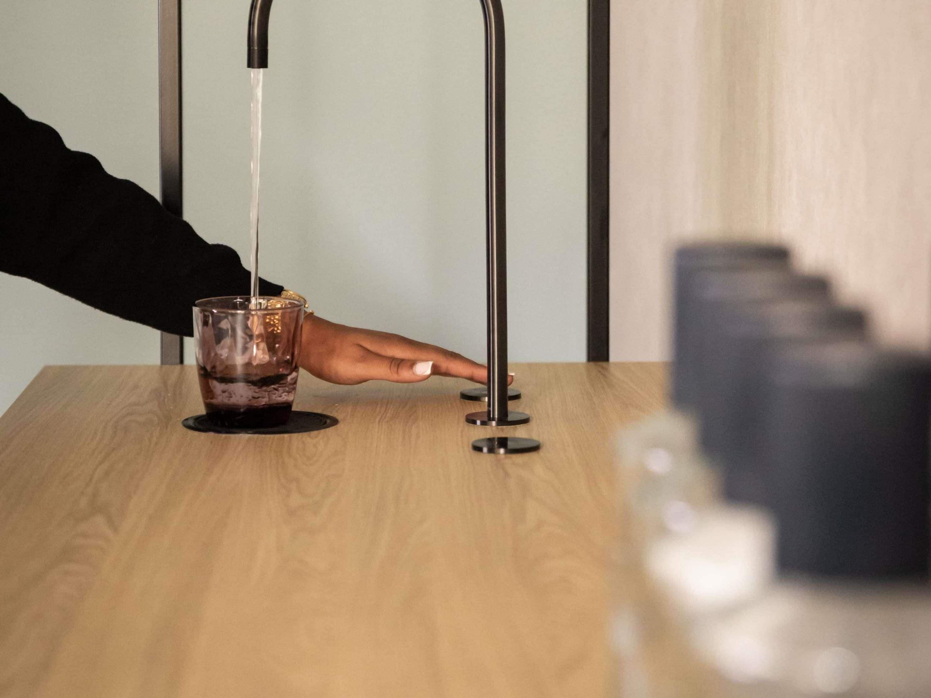 A hand pouring water into a glass on a wooden table with a modern faucet.