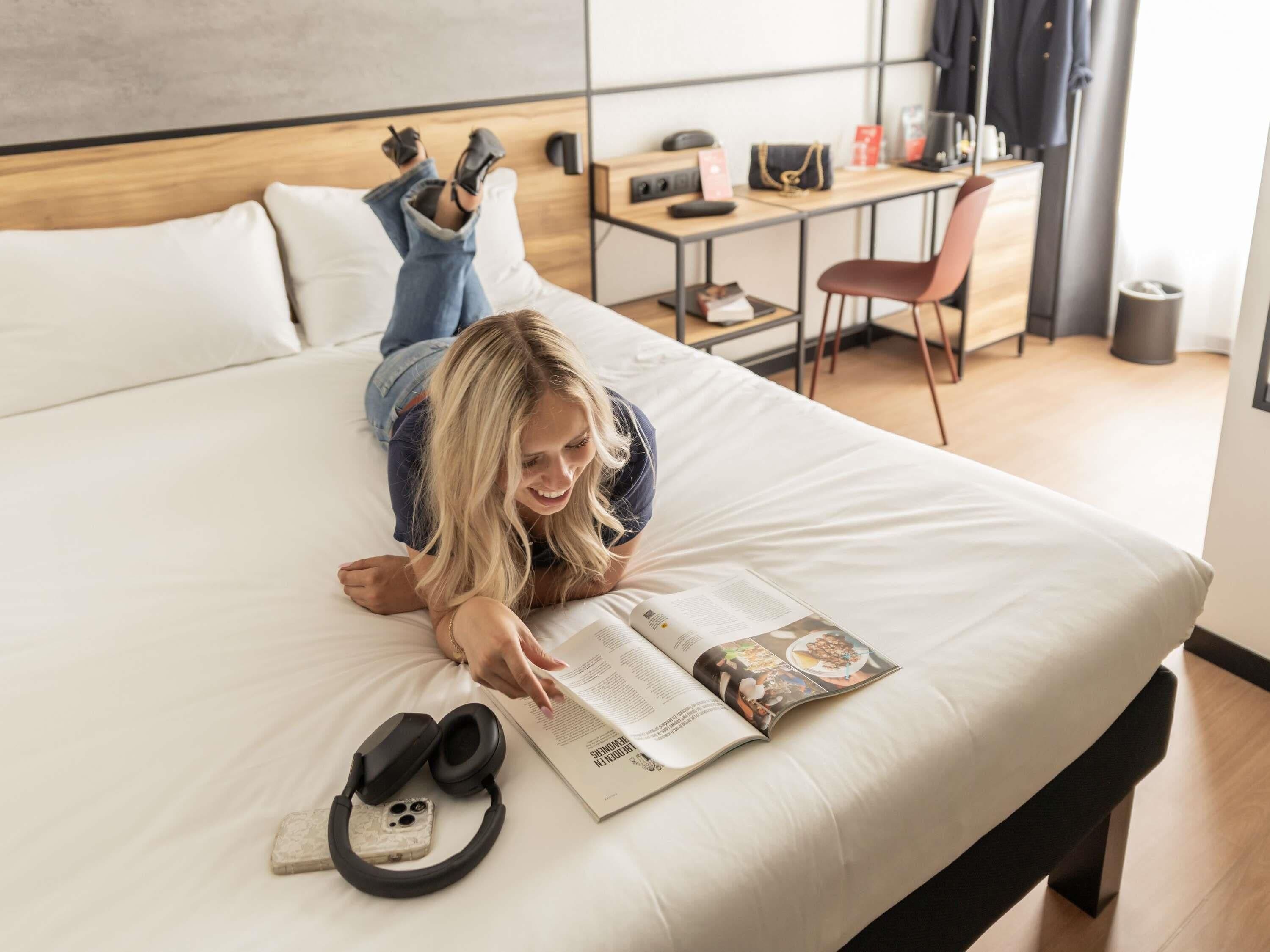 En person ligger i en seng og læser et magasin, med høretelefoner og en smartphone ved siden af.