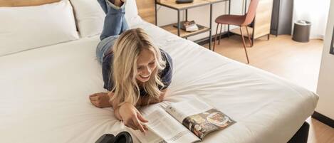 A person lying on a bed reading a magazine, with headphones and a smartphone beside them.