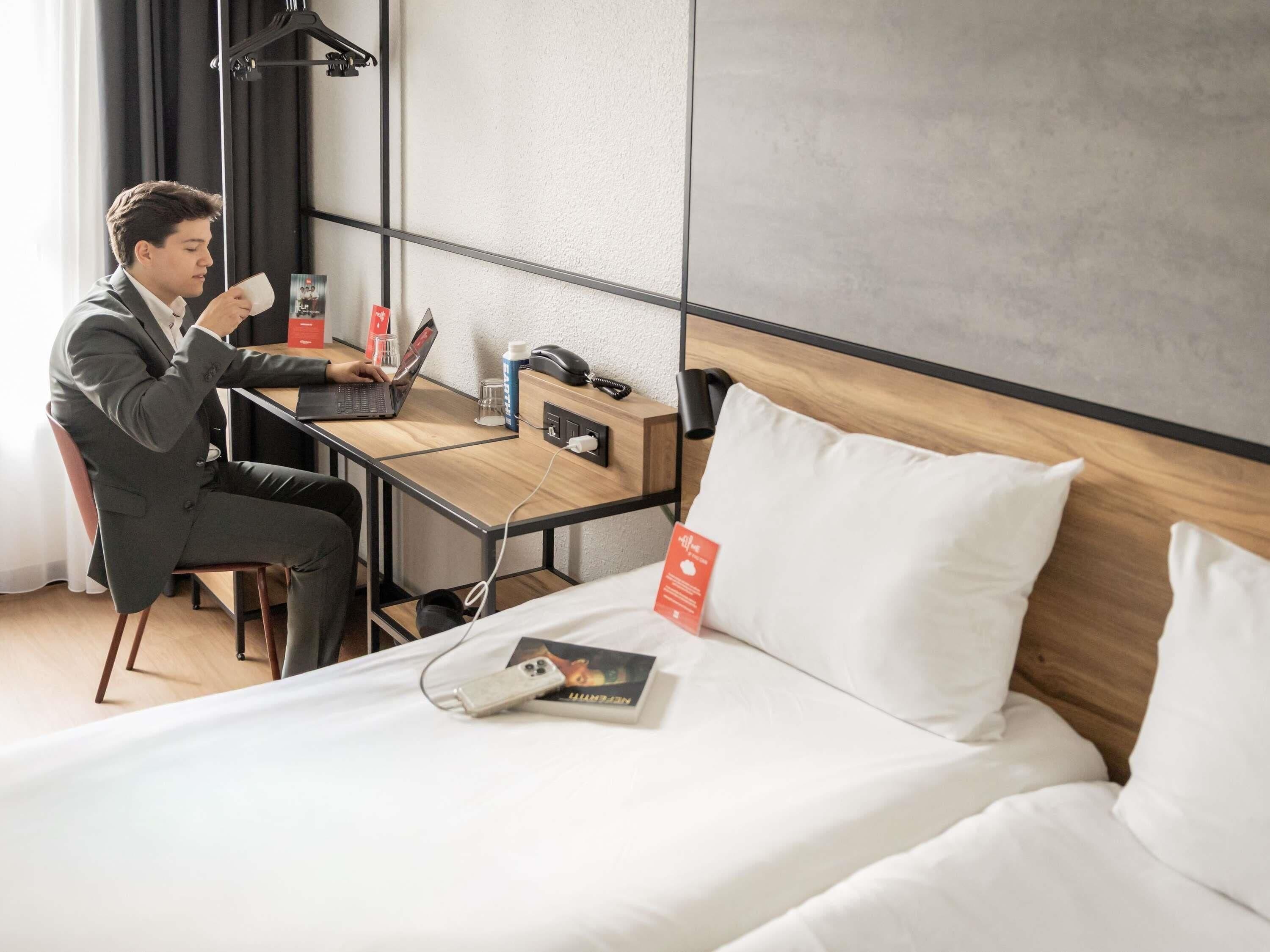 A hotel room with a bed, a desk, a laptop, a phone, and a magazine.