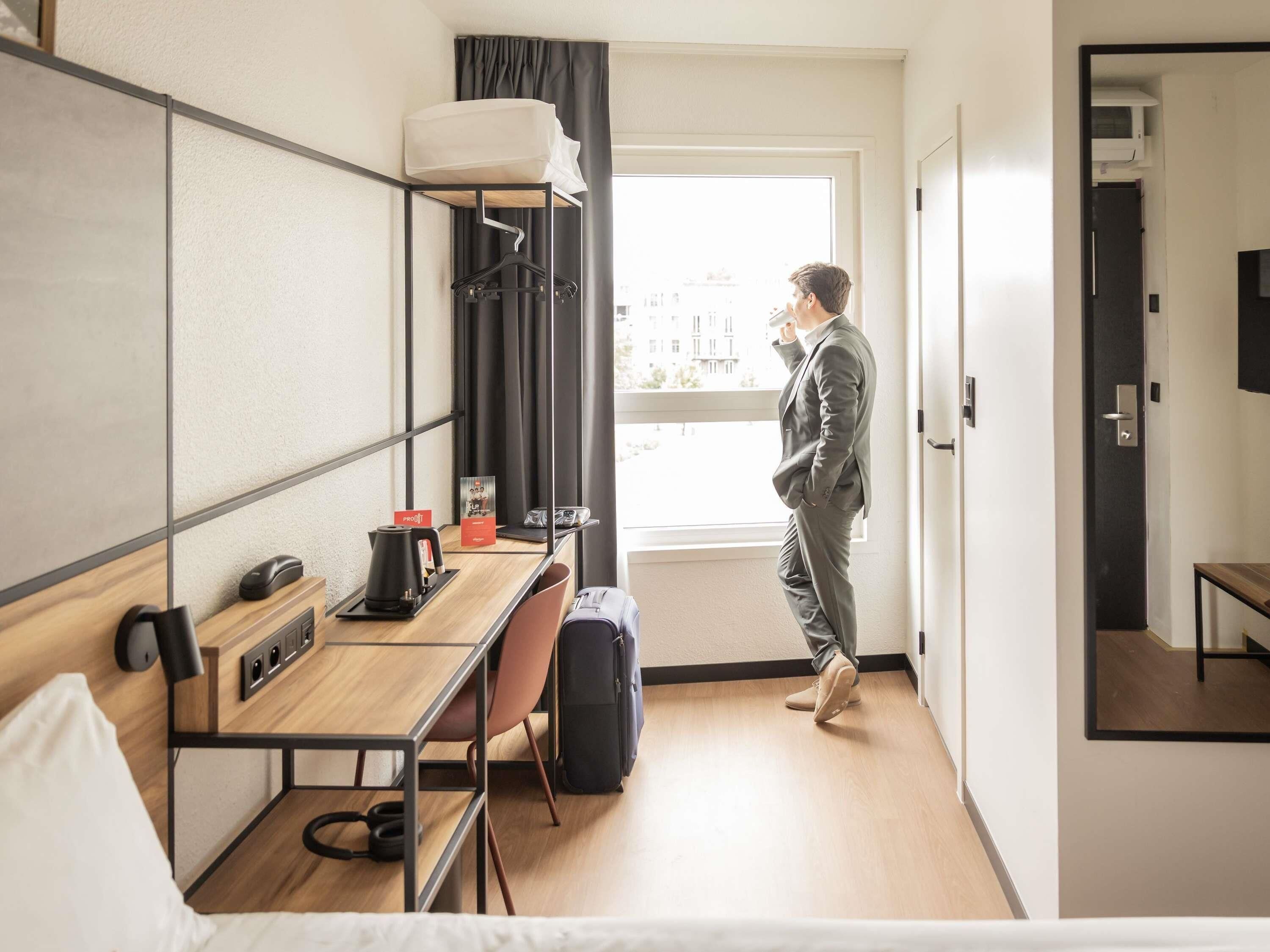 Habitació d'hotel amb un llit, un escriptori amb un ordinador, una cadira, una maleta i una finestra amb cortines.