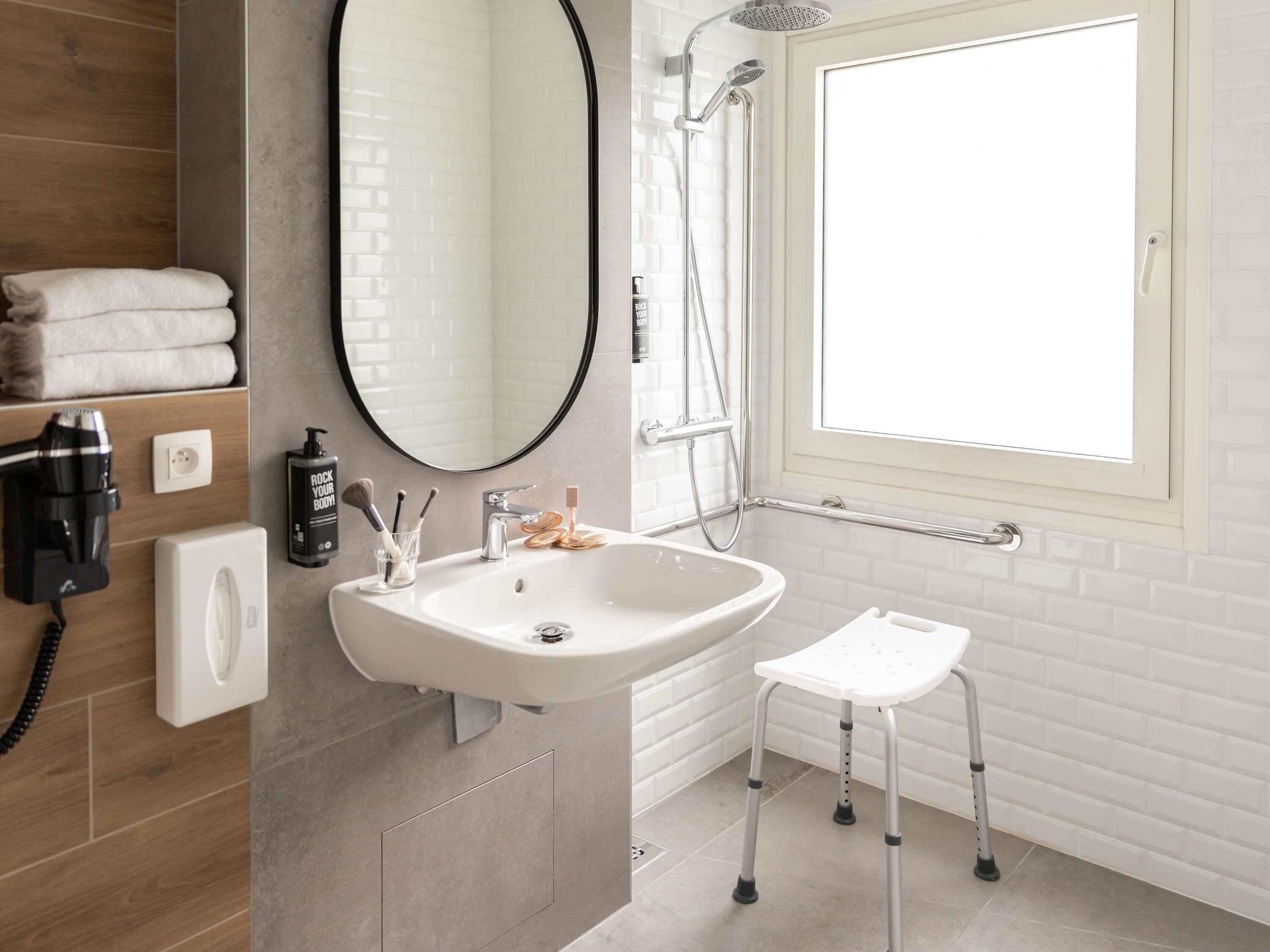 A modern bathroom with a white sink, a round mirror, a handheld showerhead, and a window.