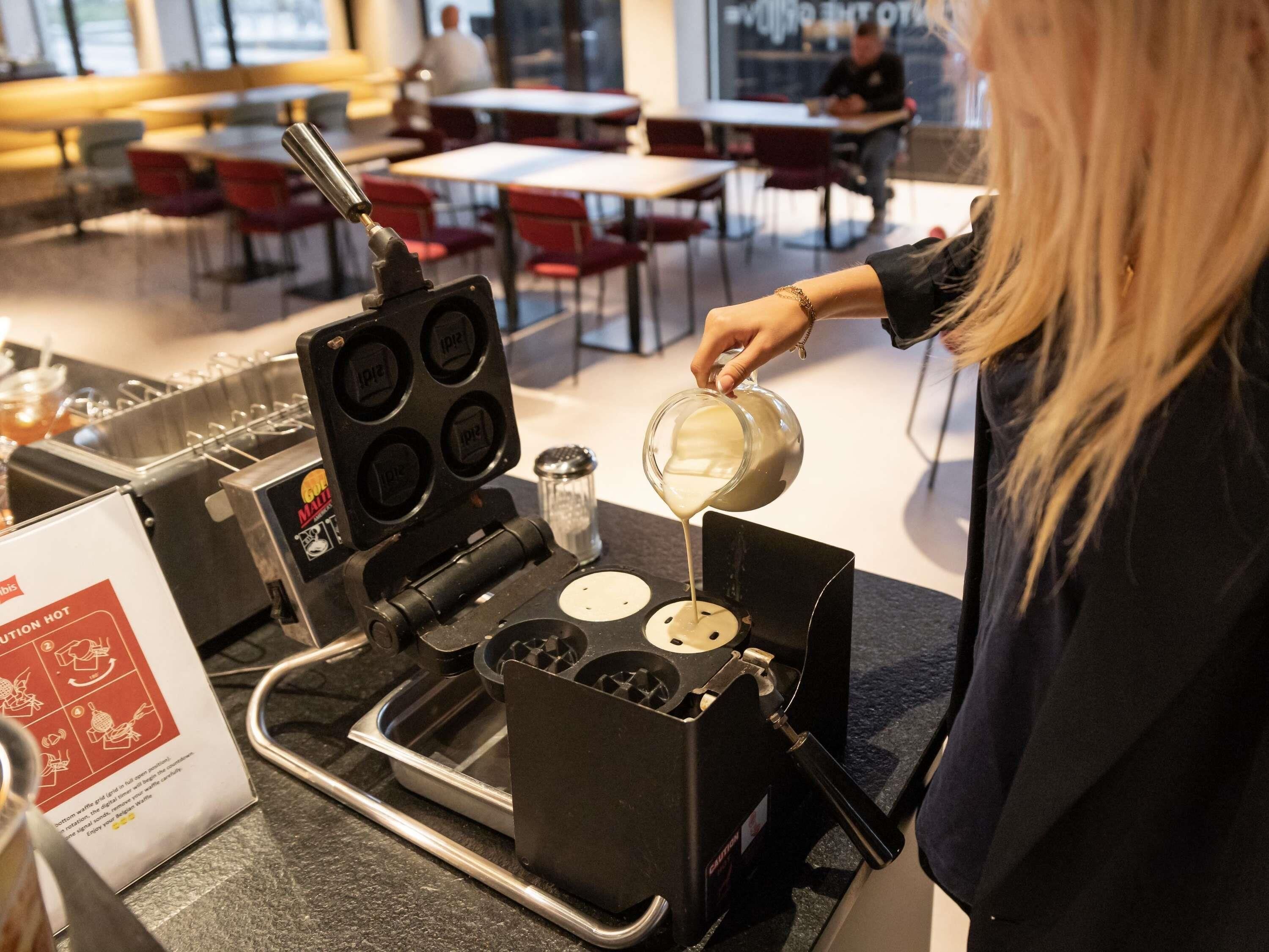 A waffle maker with multiple slots, a person pouring batter into it, and a sign with instructions.