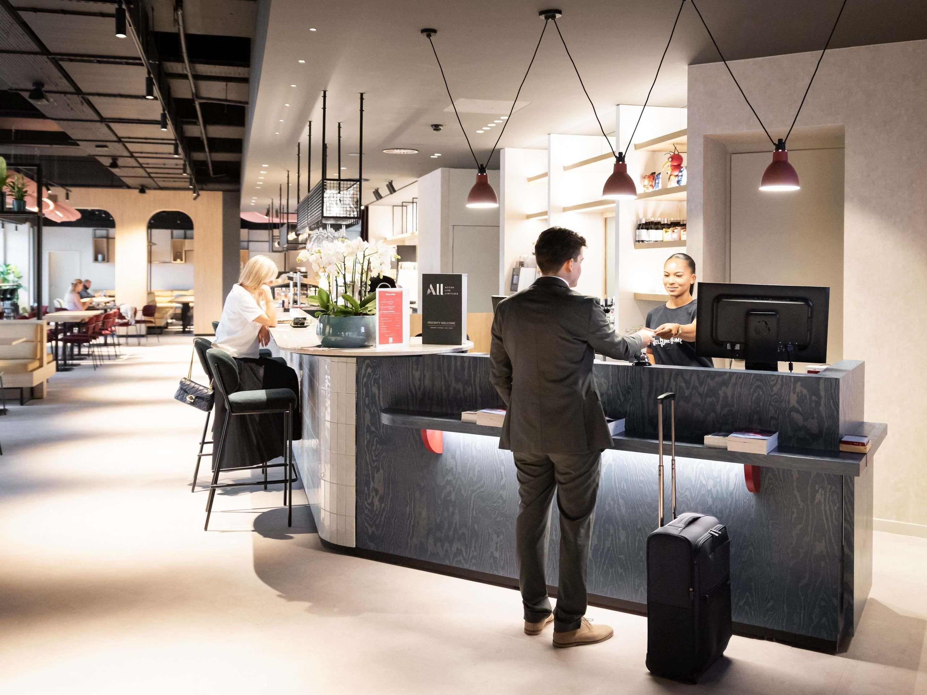 A hotel reception area with a counter, a seated guest, and a standing guest interacting with the receptionist.
