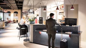 A hotel reception area with a counter, a seated guest, and a standing guest interacting with the receptionist.