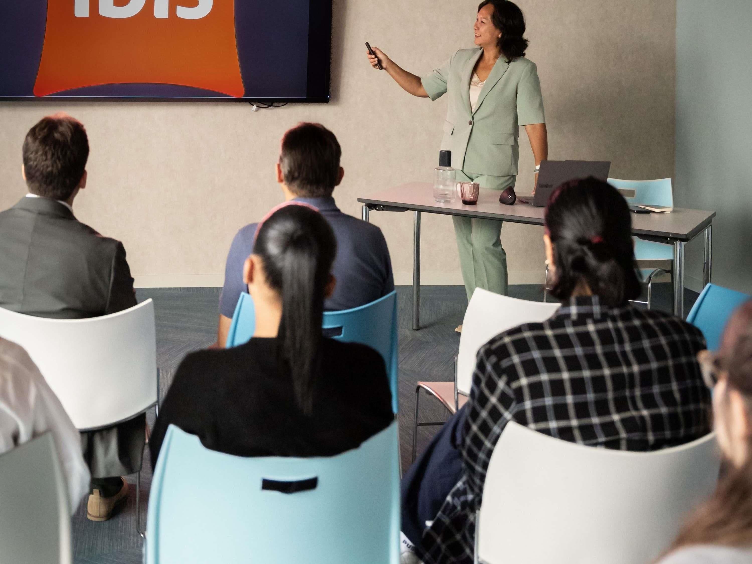 A conference room with a presentation screen, a speaker, and attendees seated facing the screen.