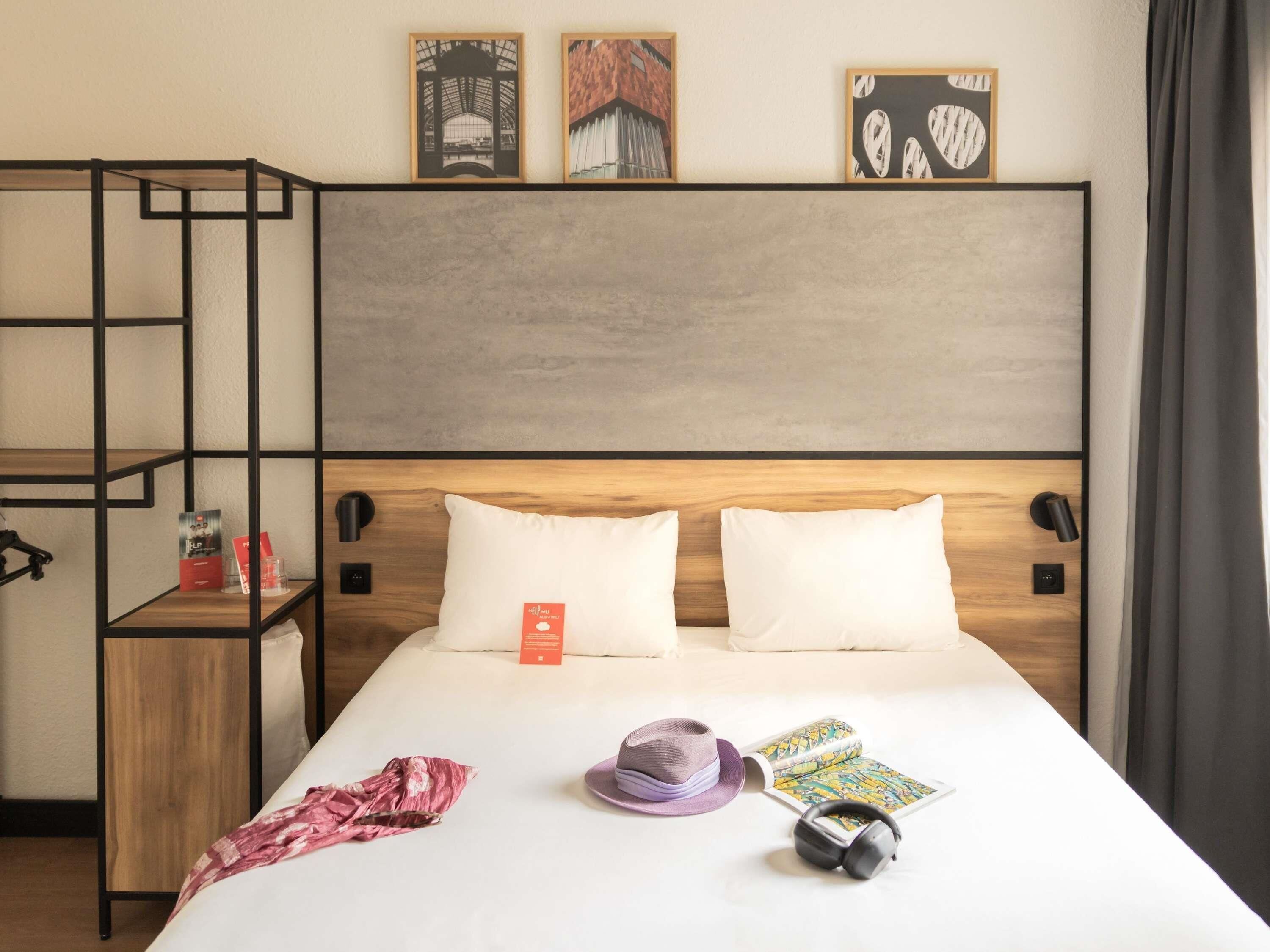 A hotel room with a bed, a wooden headboard, a shelf with books, a hat, and headphones.