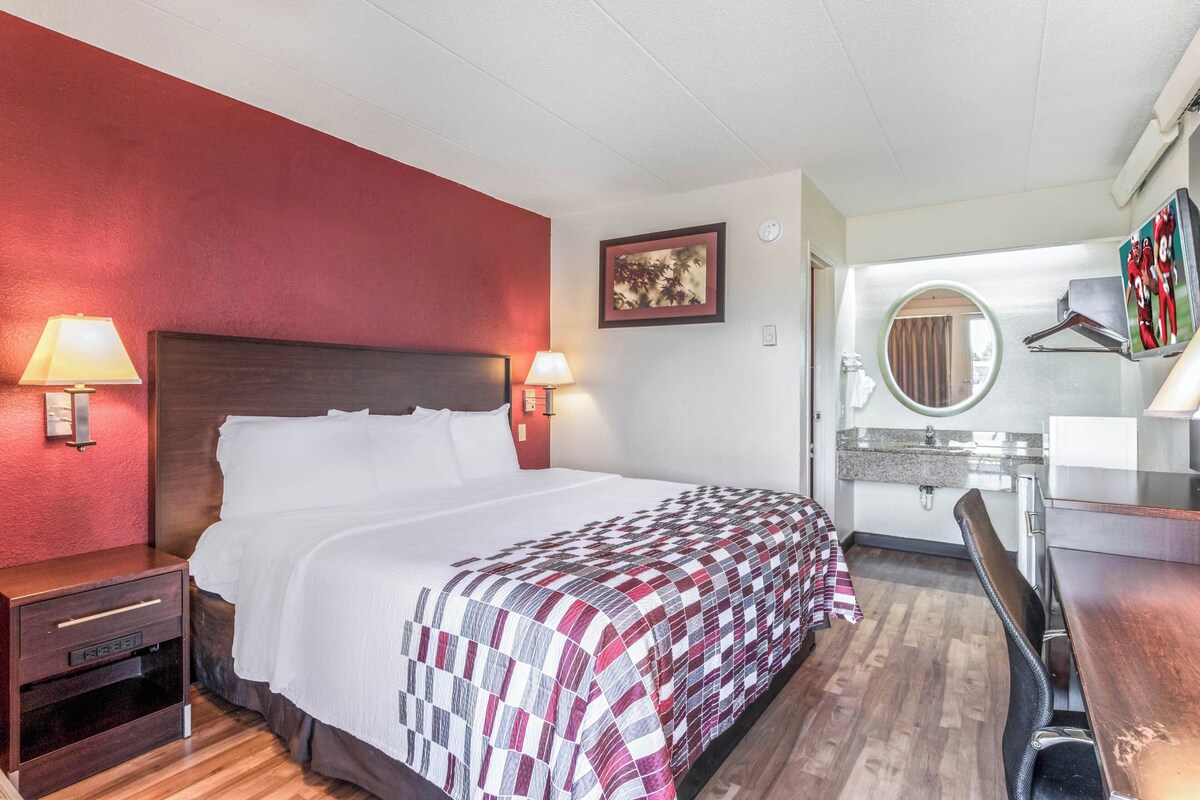 A hotel room with a bed, bedside lamps, a desk, a chair, a round mirror, and a wall-mounted TV.
