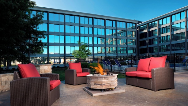 An outdoor seating area with wicker furniture and red cushions, a stone fire pit, and a modern building in the background. - Four Points by Sheraton Kansas City Airport (Kansas City)