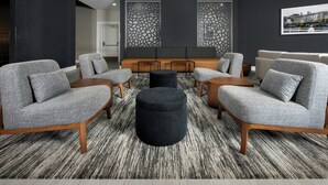 A modern lounge area with grey upholstered sofas, a central black ottoman, and a patterned carpet. - Four Points by Sheraton Kansas City Airport (Kansas City)