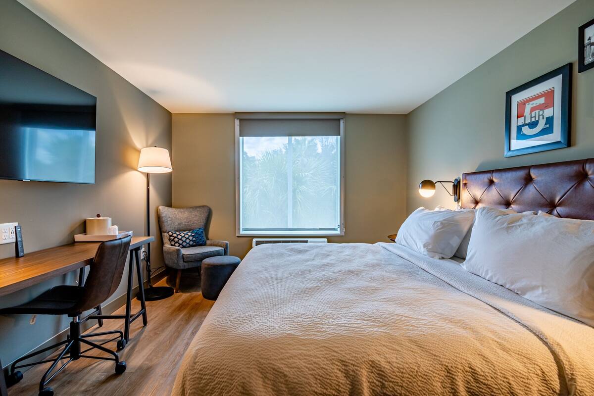 A hotel room with a bed, a desk with a chair, a TV mounted on the wall, a window with blinds, and a framed picture on the wall.