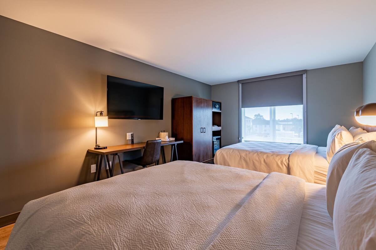 A hotel room with two beds, a desk, a chair, a TV mounted on the wall, and a window with a view.