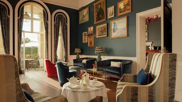 A room with a dining table set for four, surrounded by upholstered chairs. There are two sofas and a small round table with a vase of flowers. The room has a large window with curtains, and the walls are adorned with framed pictures.