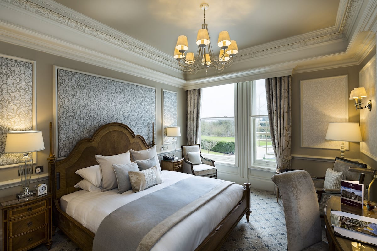 Une chambre spacieuse avec un grand lit, une tête de lit en bois, un lustre et une vue sur l’extérieur par une fenêtre.