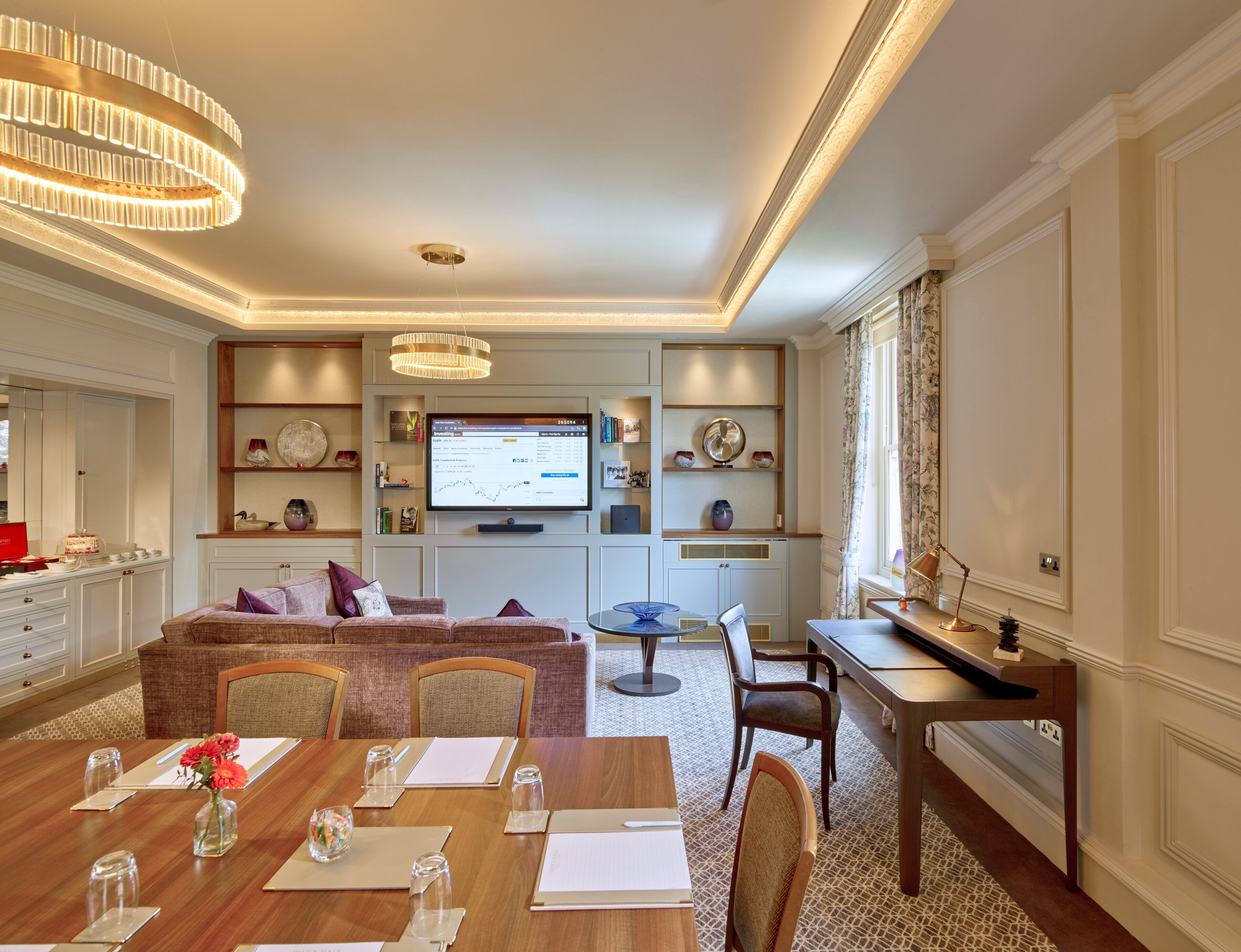 Un comedor luminoso y elegante con una mesa de madera dispuesta para una comida, una zona de estar confortable con un sofá, y un televisor que muestra un gráfico de la bolsa de valores.