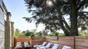 A sunlit outdoor lounge area with wooden benches and lounge chairs, surrounded by a wooden fence and a large tree.