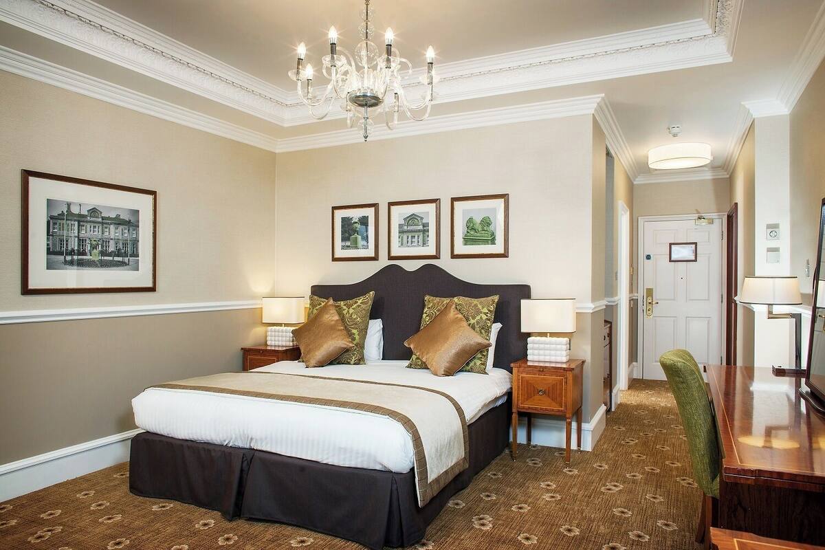 Une chambre d’hôtel avec un grand lit, deux tables de chevet, un lustre, des tableaux encadrés au mur et une porte donnant sur une autre pièce.