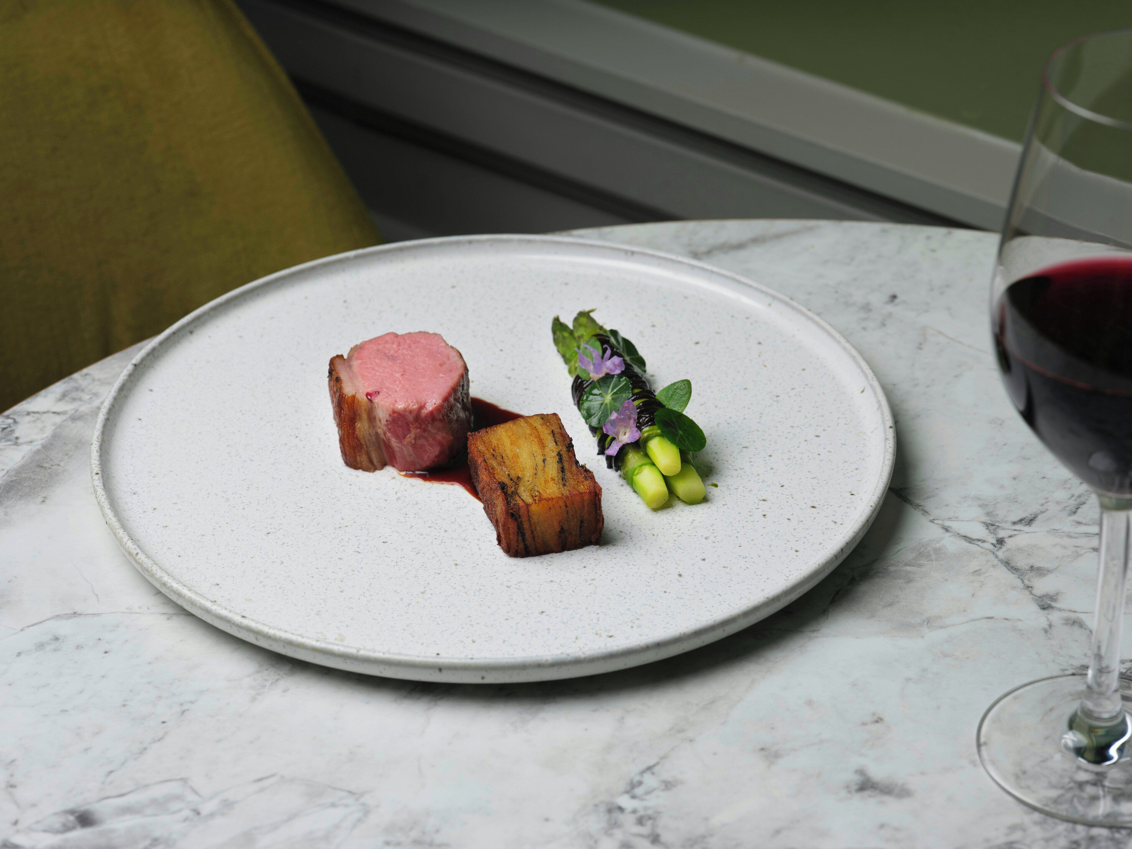 A plate with a piece of meat and vegetables on a white plate, a glass of red wine, and a green chair in the background.