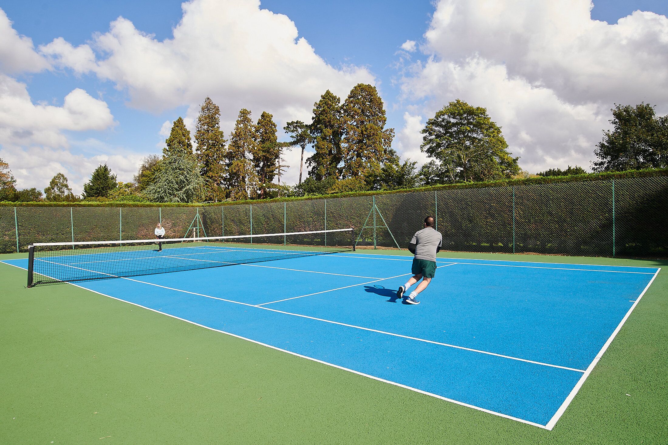 A tennis court with a net, surrounded by a fence and trees.
