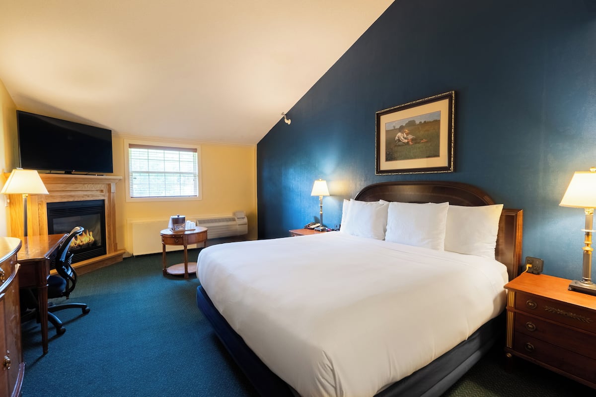 A hotel room with a large bed, a fireplace, a desk with a chair, a window, and a painting on the wall.