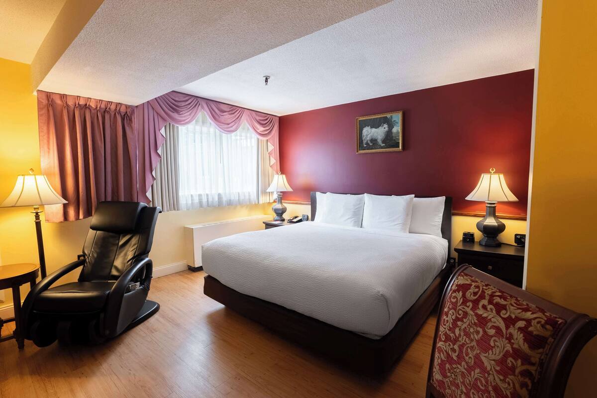 A hotel room with a bed, a massage chair, two bedside tables with lamps, a window with curtains, and a painting on the wall.