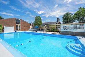 A rectangular swimming pool with clear blue water, surrounded by a white fence, a brick building with a glass door, and a covered seating area with umbrellas.