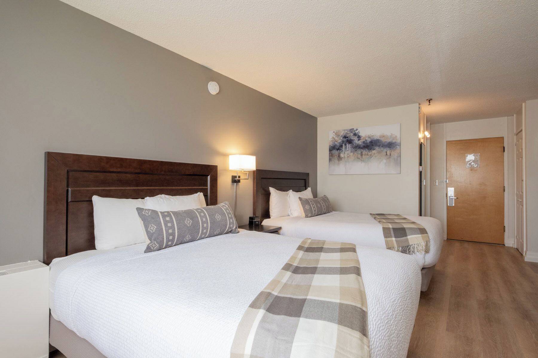 A hotel room with two beds, a nightstand, a wall-mounted lamp, a painting, and a door.
