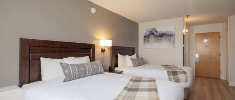 A hotel room with two beds, a nightstand, a wall-mounted lamp, a painting, and a door.