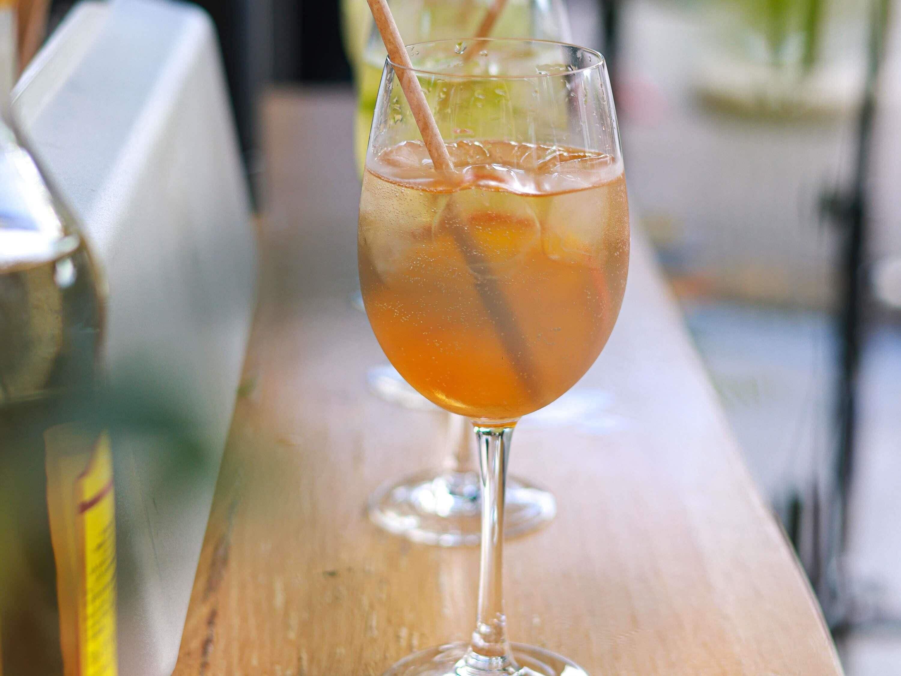 A glass of amber-colored beverage with ice cubes and a straw, placed on a wooden surface.