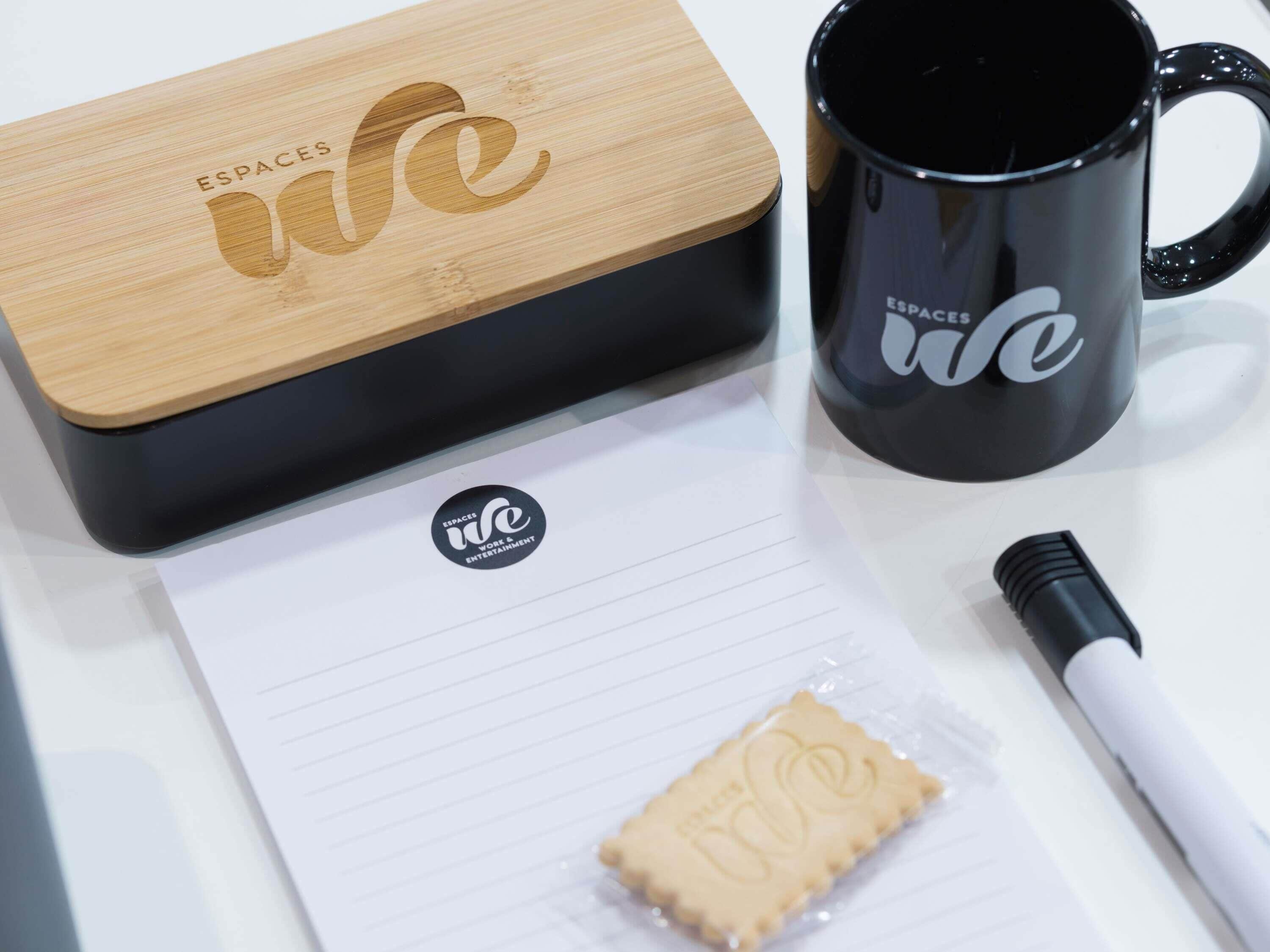 Caixa de madeira com a inscrição "ESPACE WE", uma caneca preta com a mesma inscrição, uma caneta branca e um pedaço de papel com o logotipo "ESPACE WE".