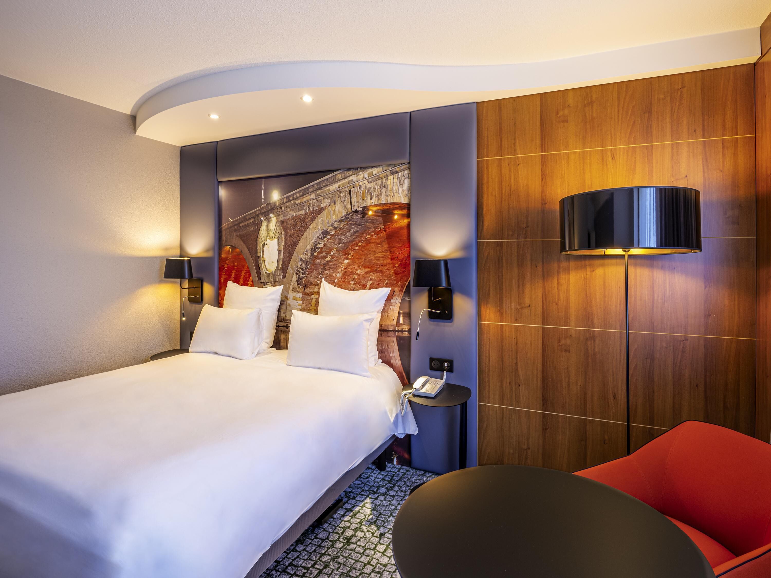 A modern hotel room with a large bed, two bedside lamps, a red chair, and a wall-mounted phone.