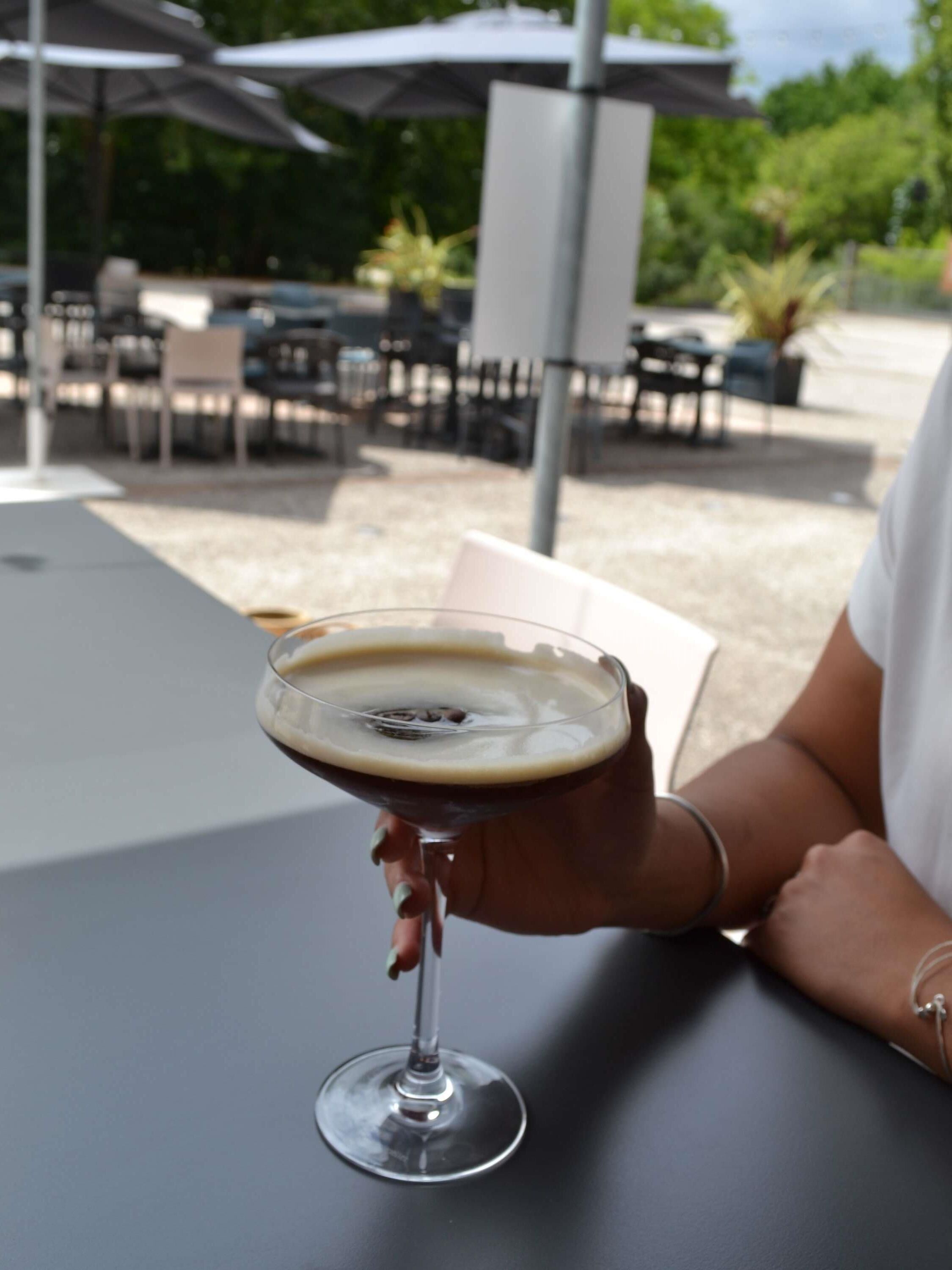 A person holding a martini glass with a dark liquid and a light foam layer, seated at an outdoor table with a white umbrella and chairs in the background.