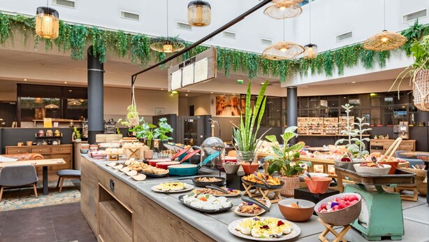 Een buffetopstelling in een moderne hotellobby met diverse gerechten, houten meubels en hangende rieten lampen.