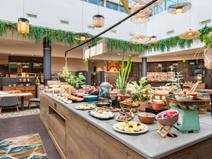 Disposição de buffet em um lobby moderno de hotel, com diversos tipos de comida, móveis de madeira e luminárias de vime pendentes.