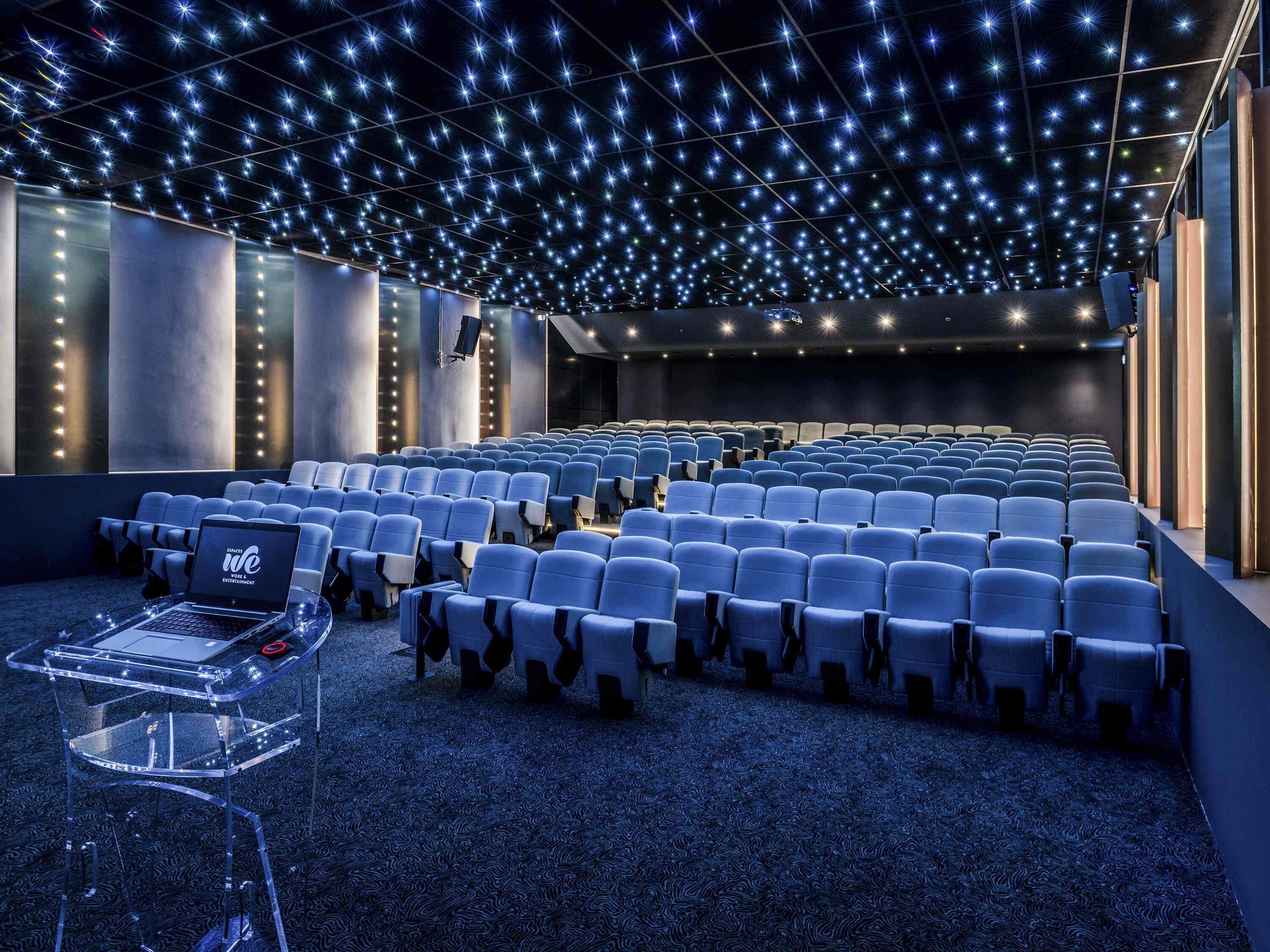 Ein modernes Kino mit Sitzreihen, einem Laptop auf einem Tisch und einer Decke mit sternförmigen Lichtern.