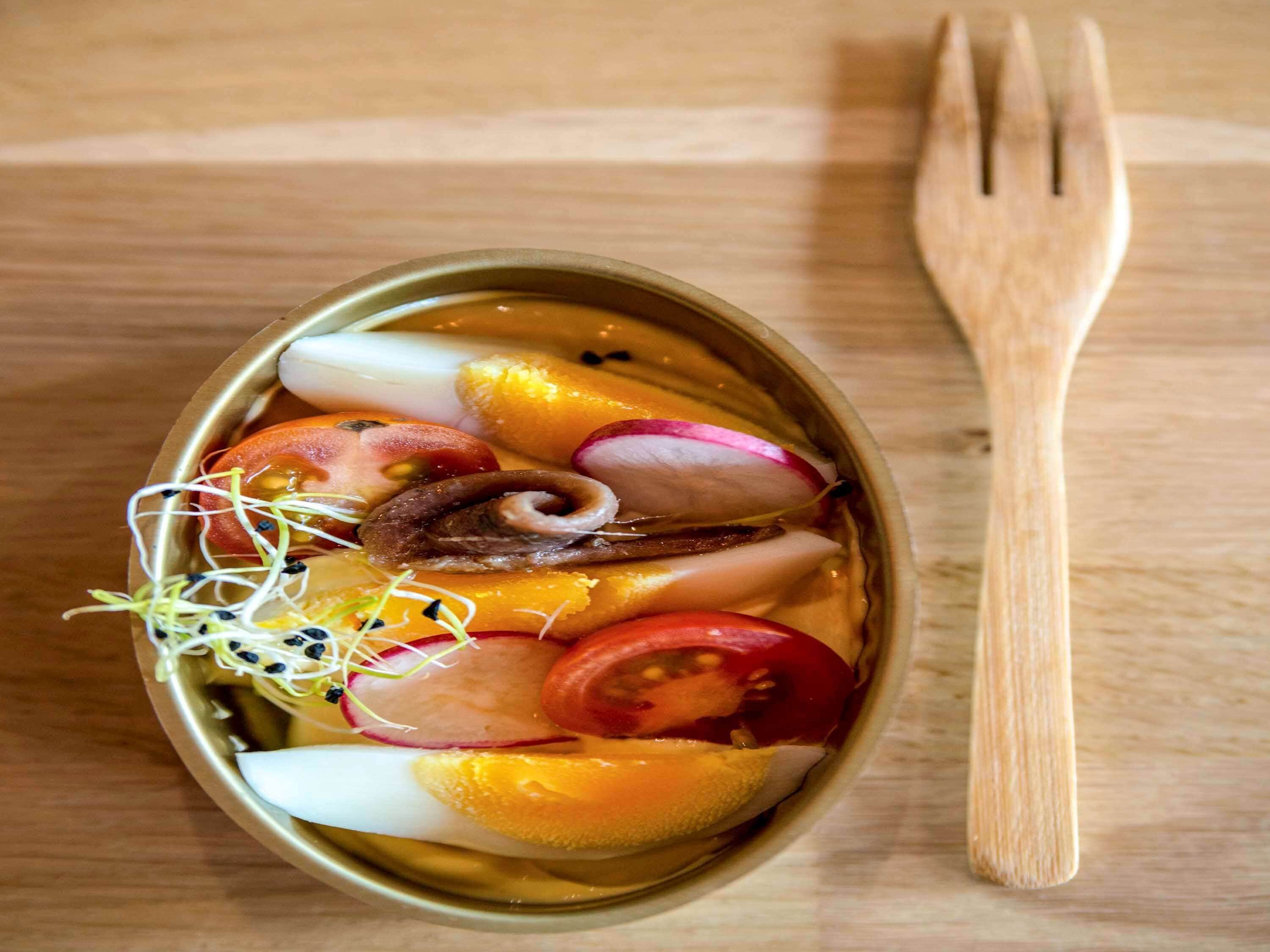 A meal in a metal bowl with various vegetables and sprouts, accompanied by a wooden fork on a wooden table.