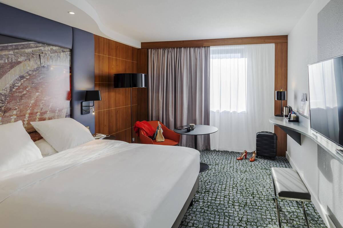 Une chambre d’hôtel avec un grand lit, une petite table avec une chaise, une télévision et une fenêtre avec des rideaux.