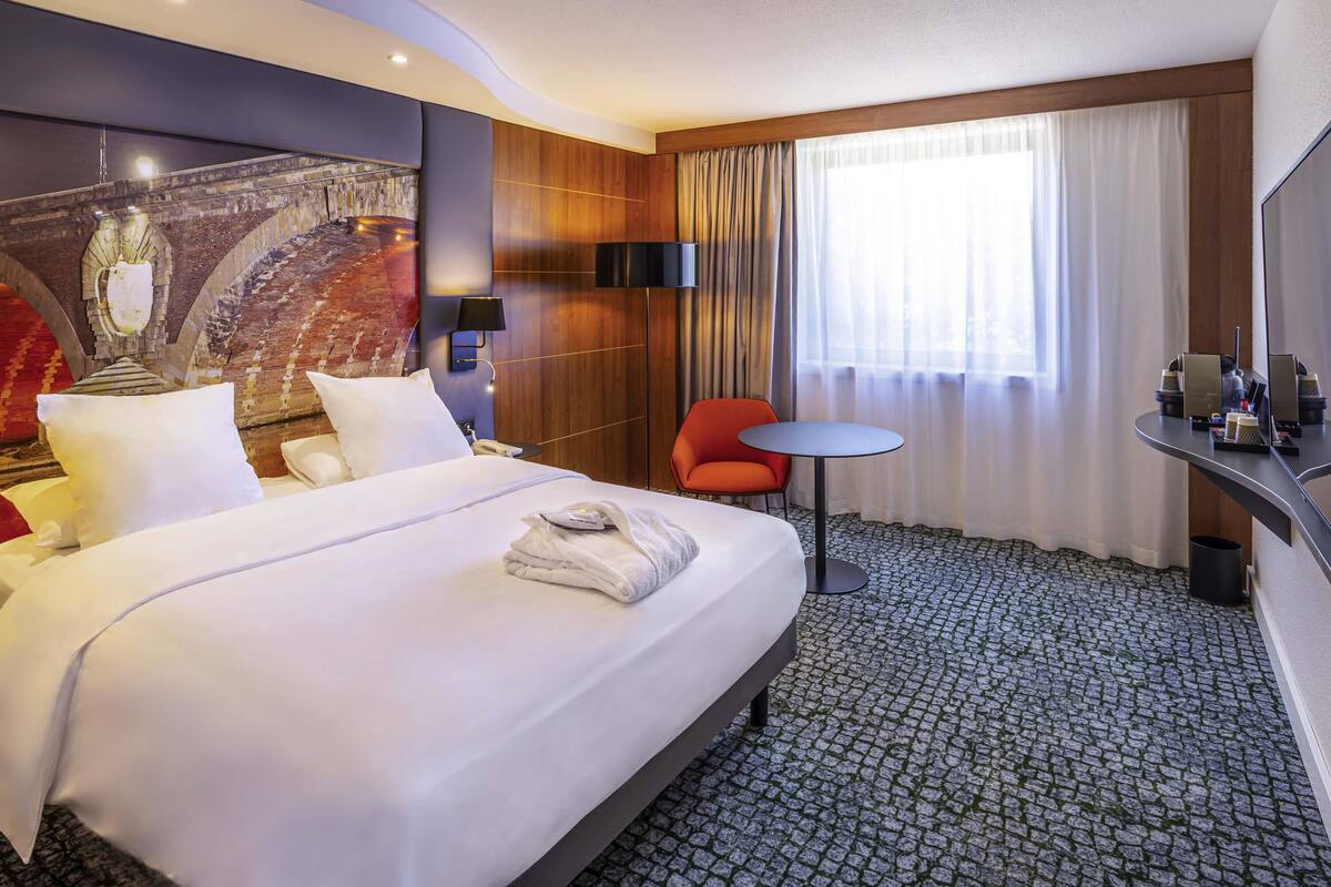 Une chambre d’hôtel avec un grand lit, une petite table ronde, une chaise rouge et une grande fenêtre avec des rideaux.